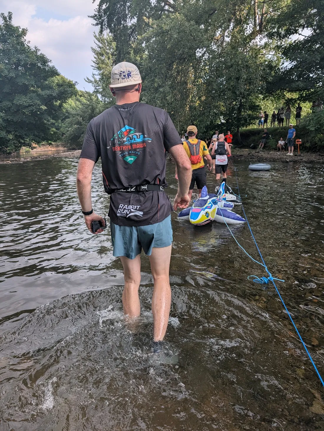James Prosser wading a river crossing during a trail race, seen from behind, with other runners ahead following a rope line